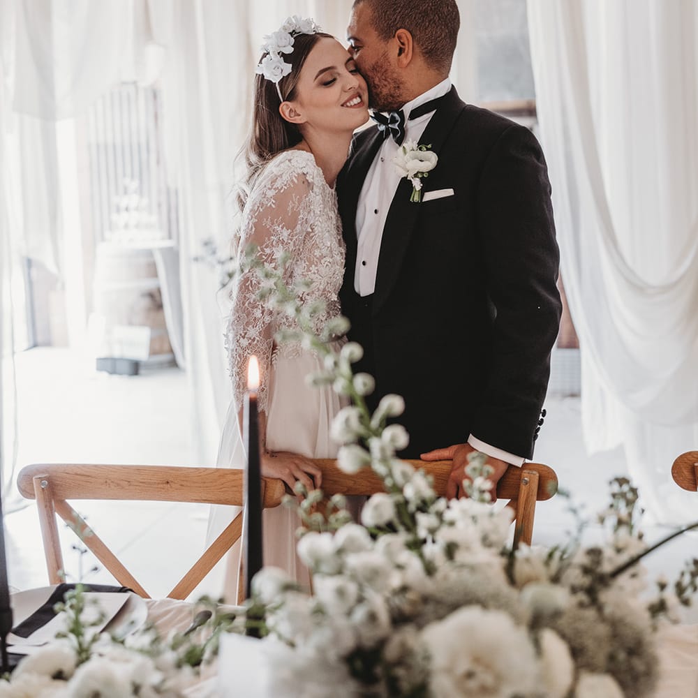 Groom in black tie kisses the bride on the cheek as they stand at their wedding reception 