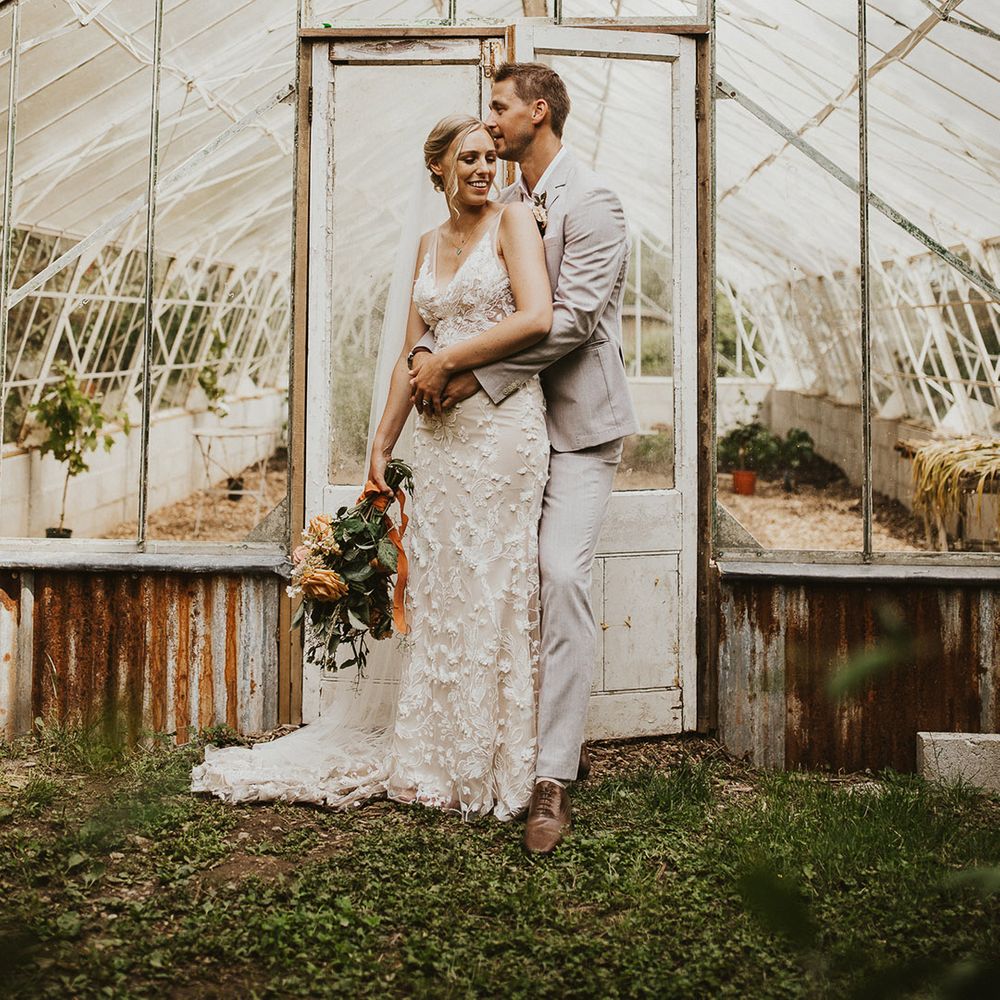 Groom in a light suit embraces the bride from behind in a lace wedding dress in front of a greenhouse at Nancarrow Farm 