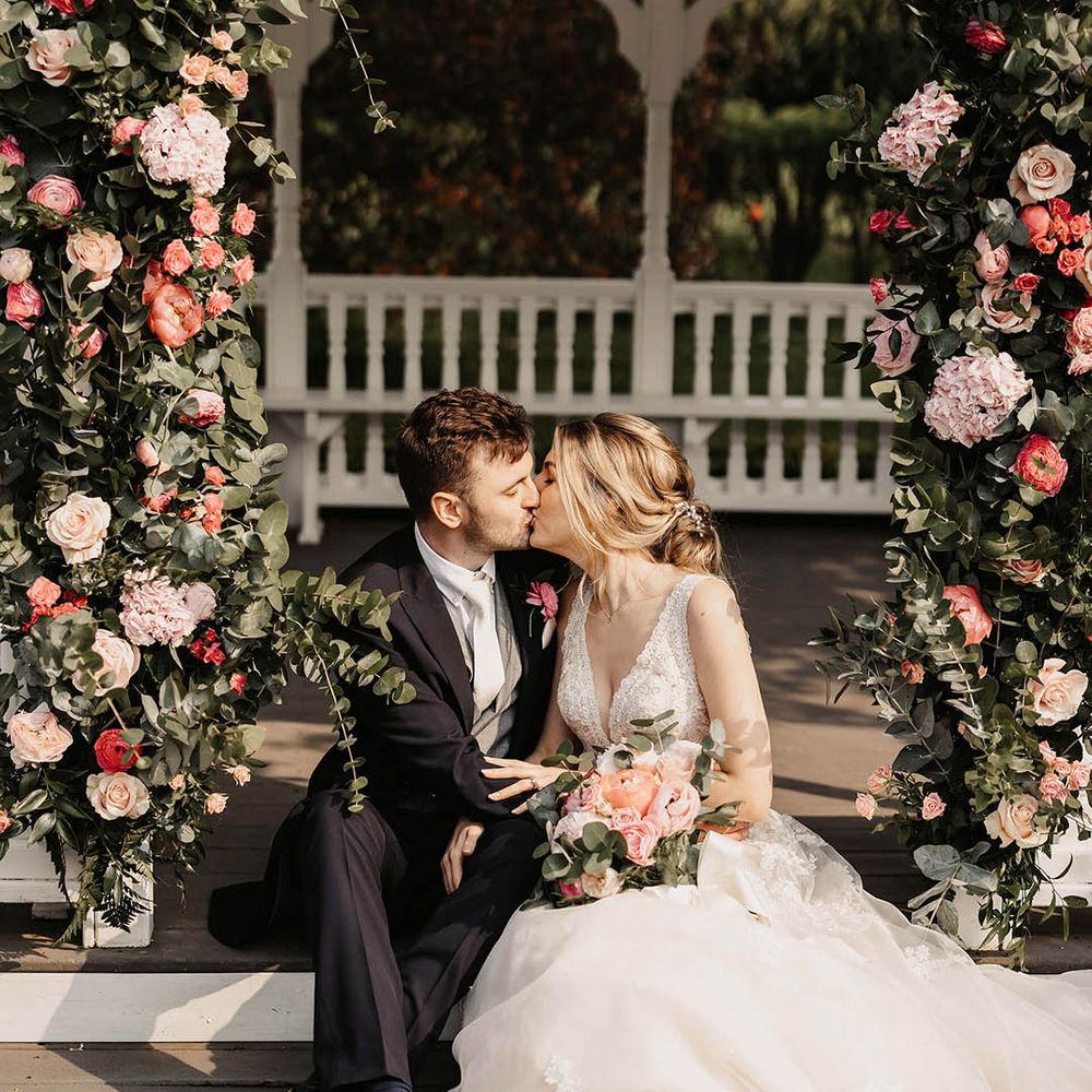 Bride and groom sit on the steps and share a kiss with a pink flower arch 