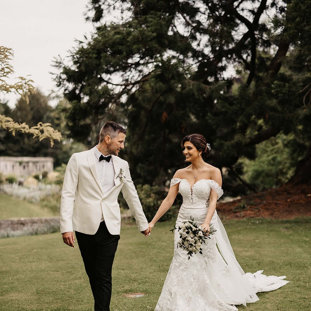 The groom in a white tuxedo walks hand in hand with the bride in a flower lace wedding dress and white flower bouquet 