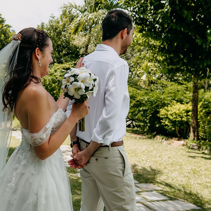 Bride in off the shoulder lace wedding dress with lace floral details holding white tropical bouquet, tapping groom in chinos and white shirt on the shoulder