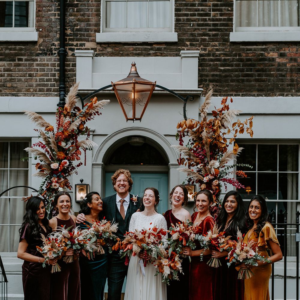 Bride in long sleeve lace vintage wedding dress and closed toe satin heels, groom in dark suit with orange tie and autumnal boutonniere and bridal party in mismatched velvet bridesmaid dresses all holding large autumnal bouquets, standing underneath large floral arch with roses, peonies, foliage and pampas grass at The Zetter Townhouse 