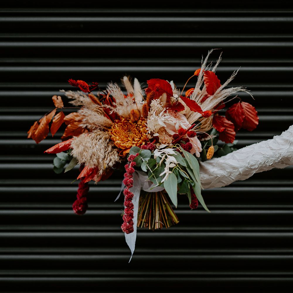 Bride holding large autumnal bouquet with orange, black, green and yellow roses, peonies, foliage and pampas grass