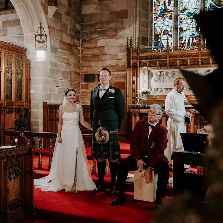 Bride in sparkly wedding dress and groom in kilt stand as the choir plays 