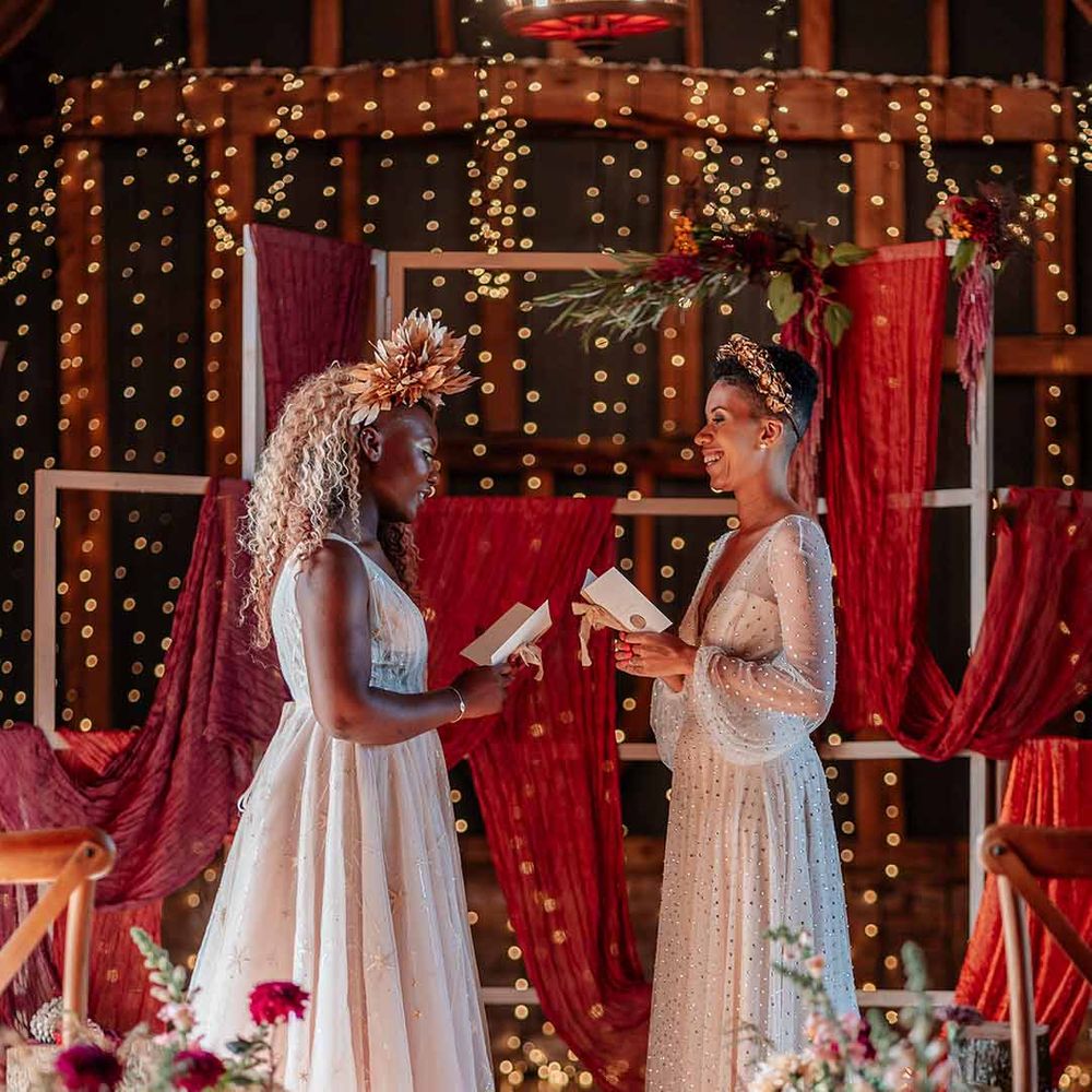Bride in sleeveless floaty peach gown and gold leaf crown and bride in crystal embellished gown and gold headdress reading each other vows at the alter 