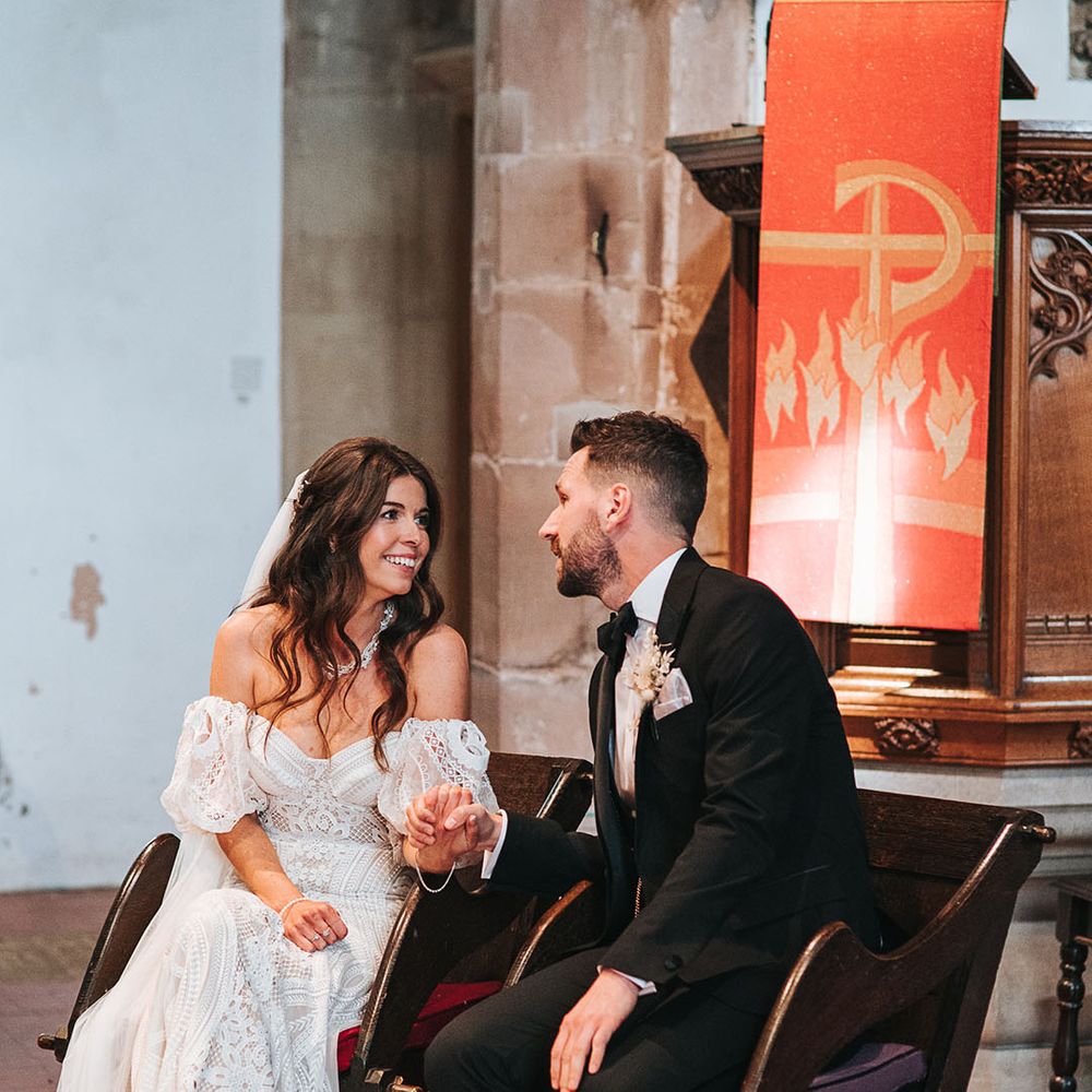 Groom in a black tuxedo with dried flower buttonhole sitting with the bride for the church ceremony in a lace wedding dress with detachable sleeves 