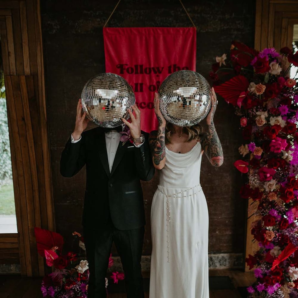 Groom in classic black tuxedo with magenta amaryllis and orchid boutonniere and bride in cowl neck sleeveless satin wedding dress with gold glittery wedding headband, and soft grey fluffy feather open toe strappy high heels, both holding up disco ball wedding decorations in front of deep pink fabric wedding banner