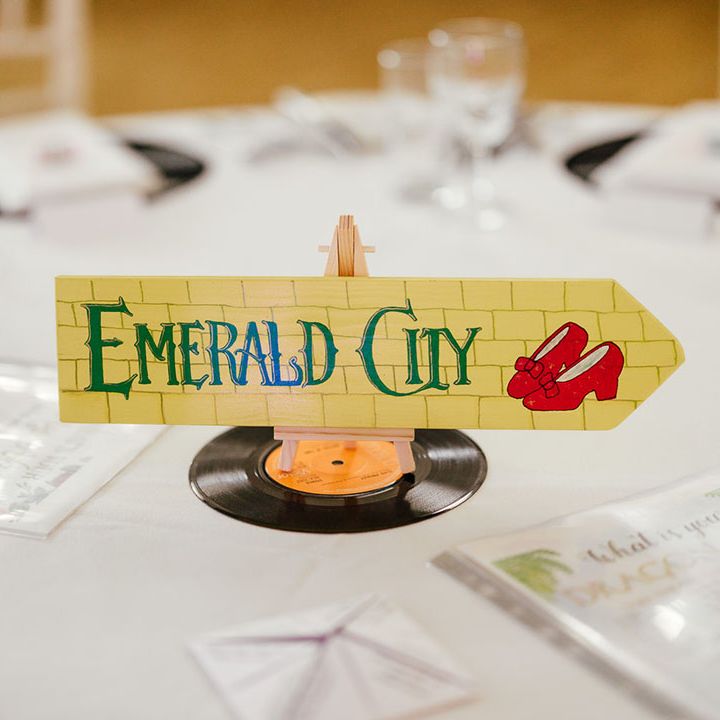 Film inspired wedding table number in the style of The Wizard Of Oz; a yellow brick patterned arrow sign with ruby red slippers and "Emerald City" in green lettering 