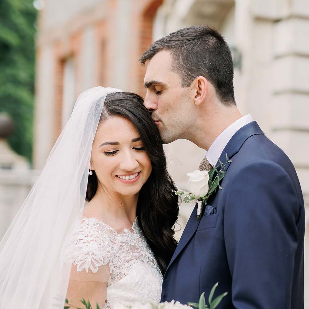 Groom in a navy suit kissing his bride's head in a fitted wedding dress with lace illusion neckline 