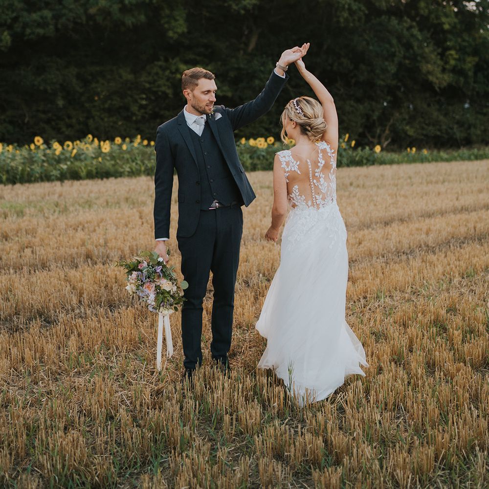 The groom in a three piece navy wedding suit spins the bride around in the fields surrounding Dove Barn Weddings 