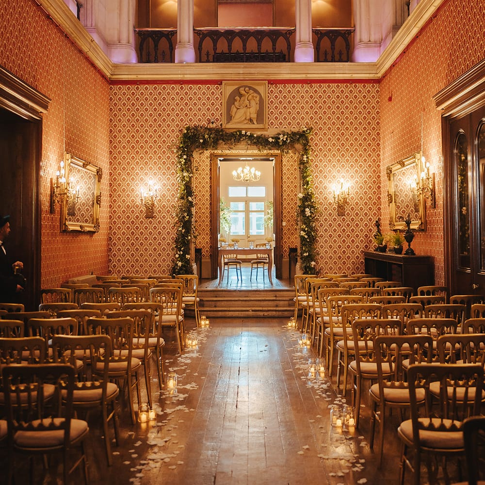 Aisle of Grittleton House wedding venue with high columns and ceilings, wooden chairs, warm cosy lighting and slim flower arch going around the door 
