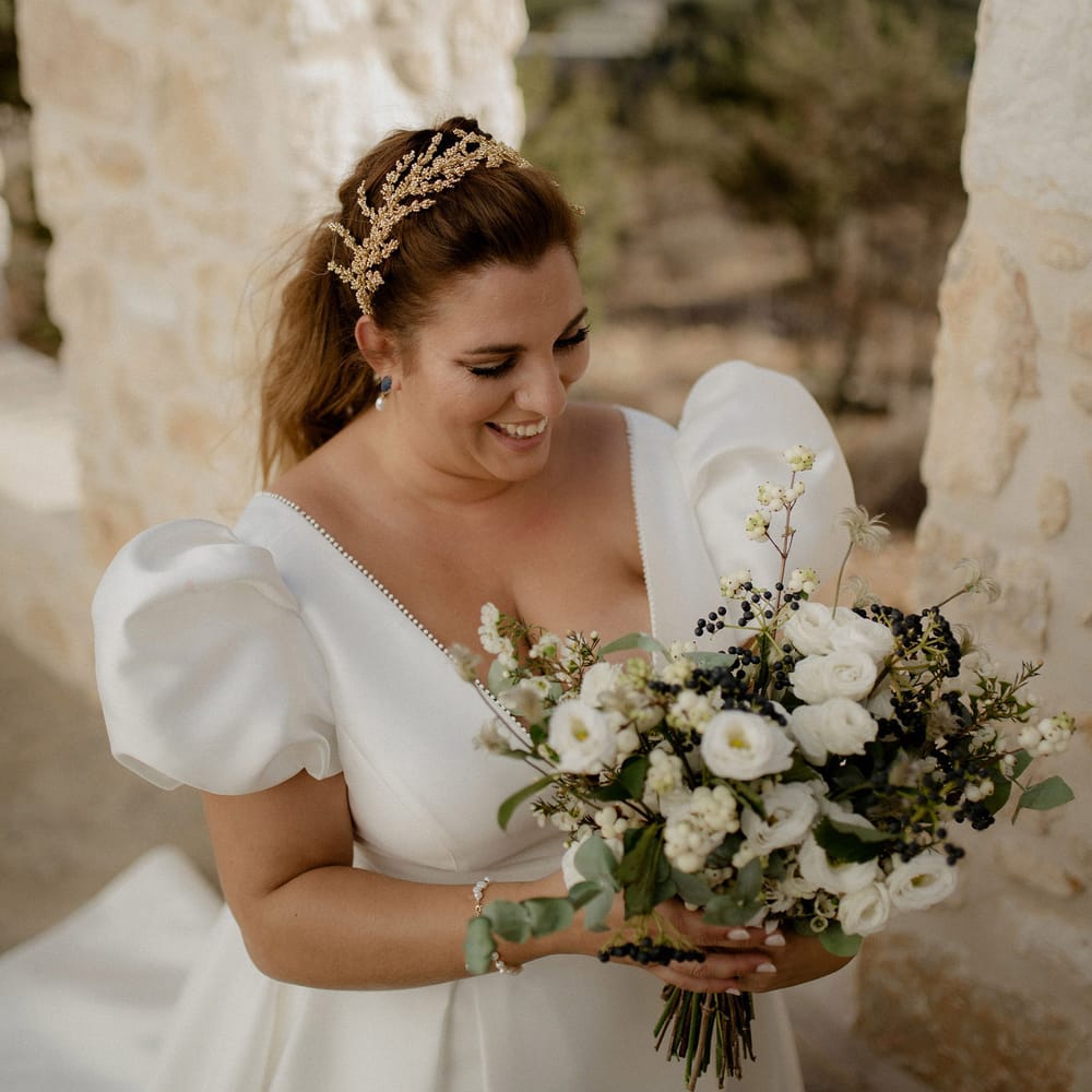 Bride carrying bridal bouqet that features wihte berries, italian ranunculus and eucalyptus whilst wearing a stain puff sleeved wedding dress, peal earrings and gold leaf bridal grown