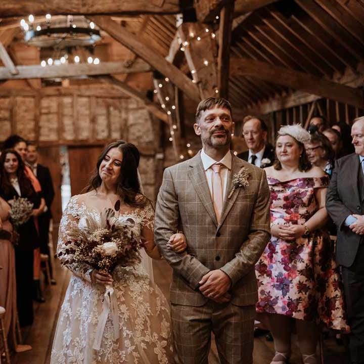 Bride in long sleeve lace illusion sleeve wedding dress with overlay at the end of the alter of Bassmead Manor Barns seeing bride in maroon bridal suit with grey waistcoat, dusky pink tie and autumnal dried flower boutonniere 