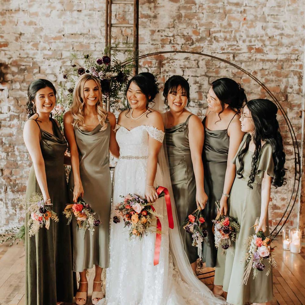 Bride in off the shoulder wedding dress standing with bridal party in mismatched satin green bridesmaid dresses 
