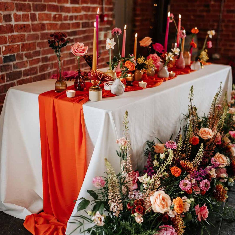 Retro wedding tablescape with pink tapered candles, orange table runner, mismatched single flower vases, colourful flower centrepieces and large colourful flower arrangements Northern Monk Refectory Leeds