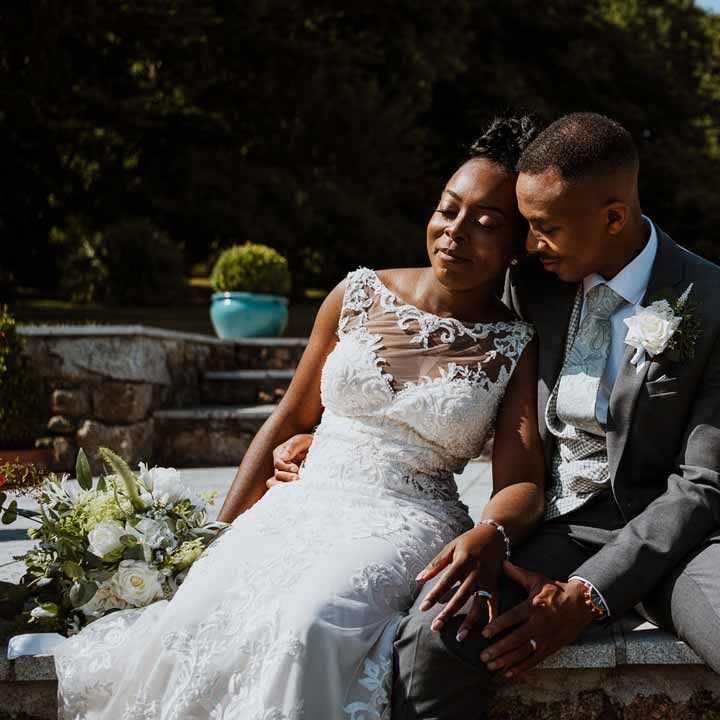 Bride in lace and mesh boatneck wedding dress with silver bridal hair accessories holding large white rose and foliage bridal bouquet sitting with groom in charcoal grey grooms suit, light grey patterned waistcoat, light grey patterned tie and large white rose and foliage boutonniere at Tredudwell Manor