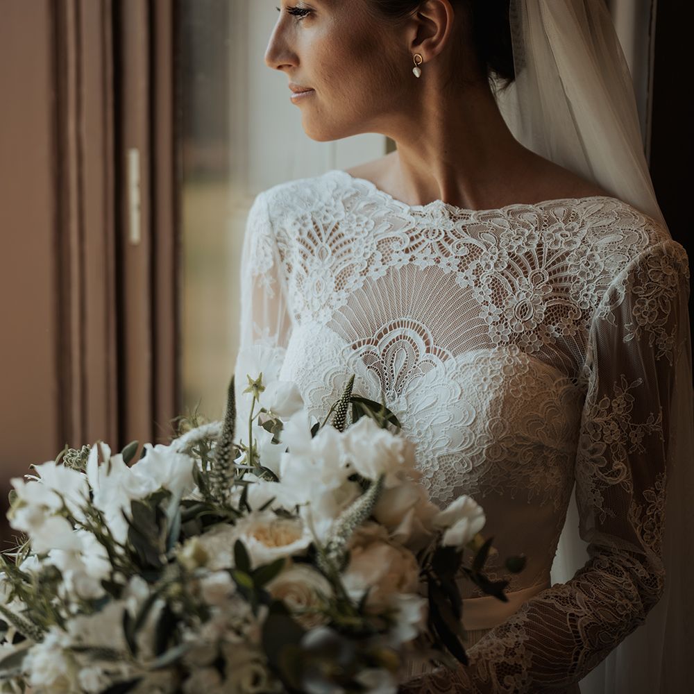bride in a long sleeve lace wedding dress looking out the window holding a white and green wedding bouquet 