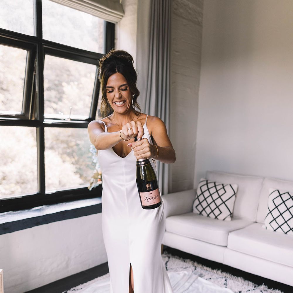 Bride pops bottle of champagne in white satin slip wedding dress 