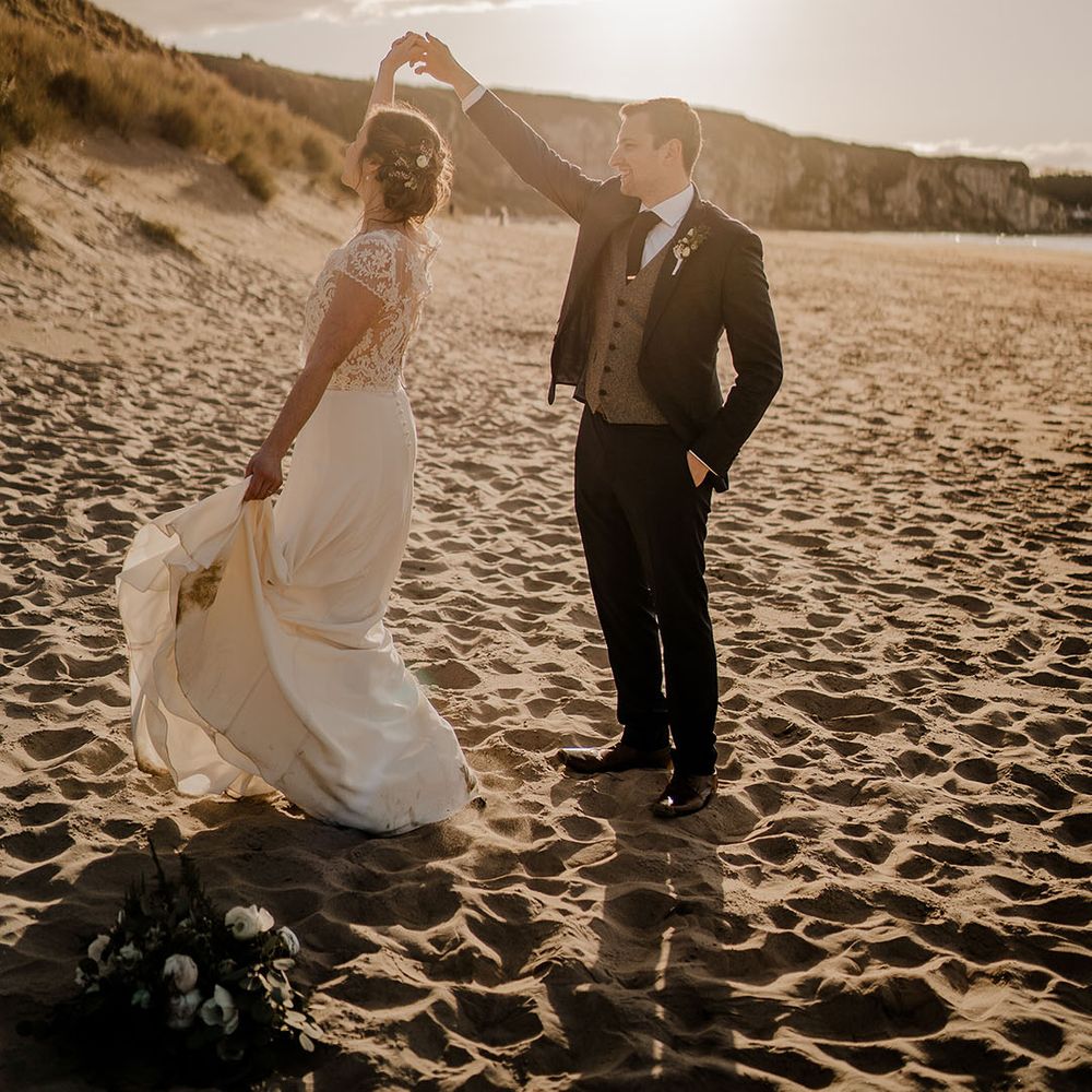 Groom in three piece suit spins the bride around at the beach on their wedding day 