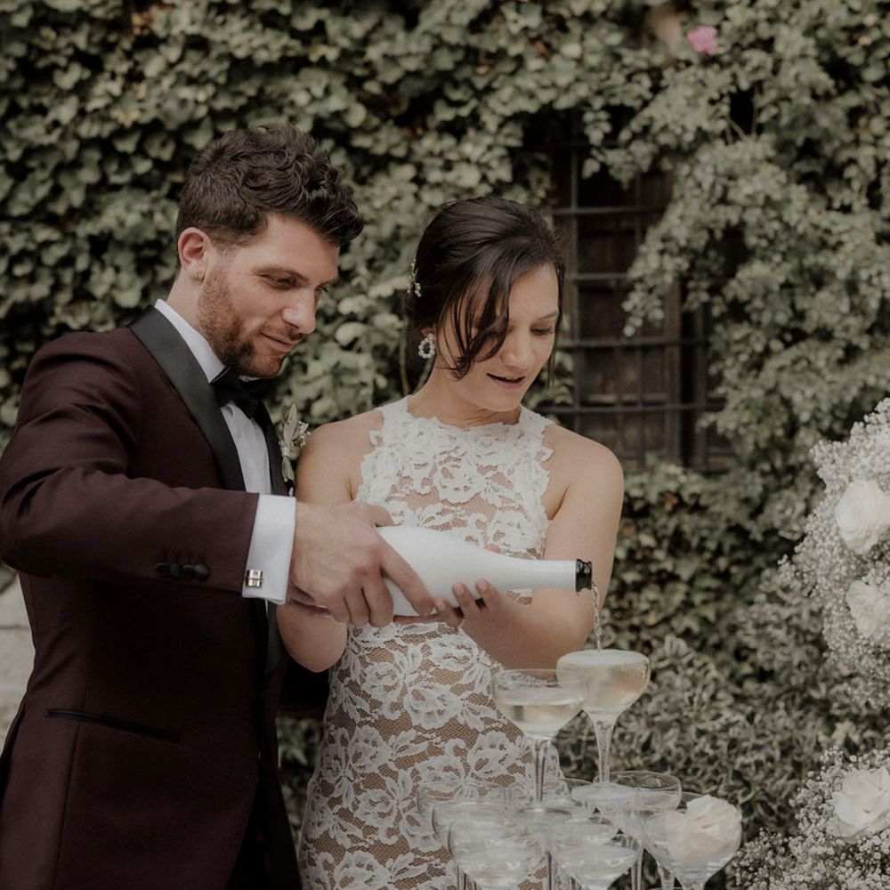 Groom in burgundy suit with bride in long sleeve lace wedding dress pouring champagne onto champagne tower 