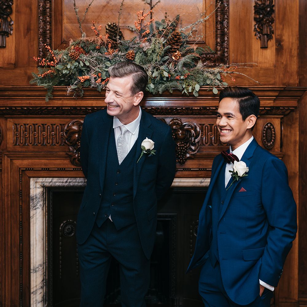 Grooms laugh and smile in front of wooden fireplace with floral decor
