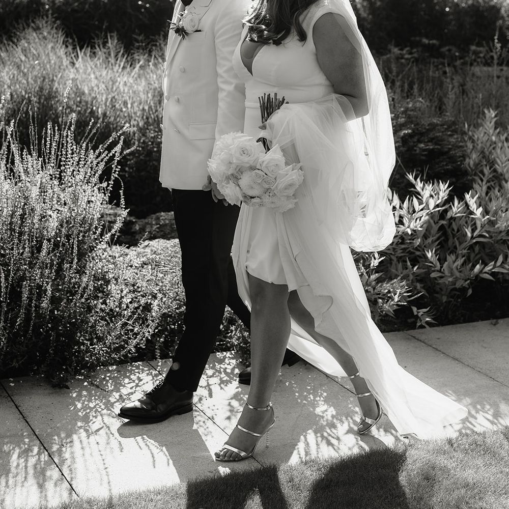 Bride in V neck Made With Love wedding dress and veil holding white rose bouquet walks outside with groom in white double breasted tuxedo jacket