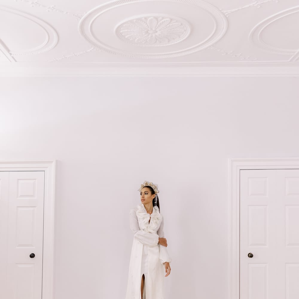 Bride stands in bright white room whilst wearing bridal gown with ruffled front and leg split