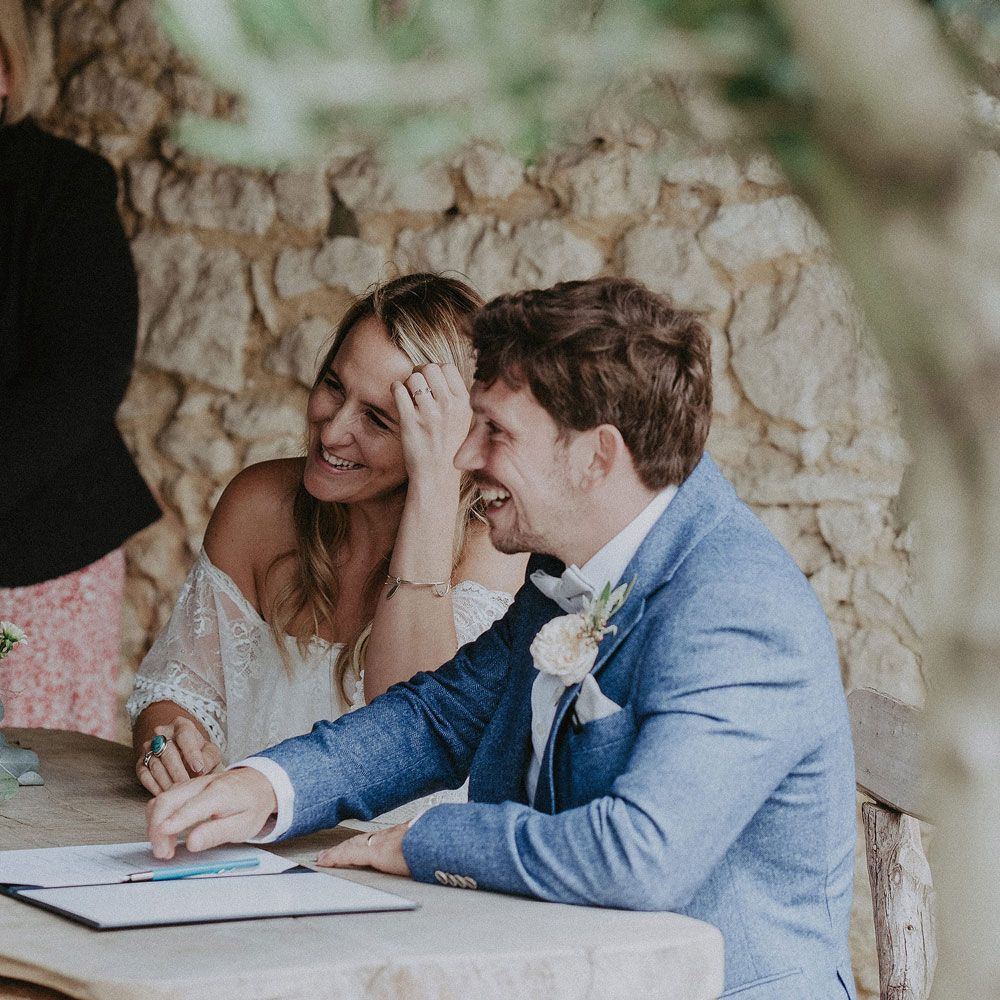 Bride in Grace Loves Lace Bardot wedding dress sits signing marriage registry with groom in blue suit with floral buttonhole at Isle of Wight wedding with macrame wedding decor