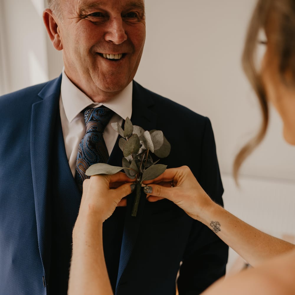 Bride applies floral buttonhole on father's blazer on her wedding day as he smiles | Mark Bamforth Photography