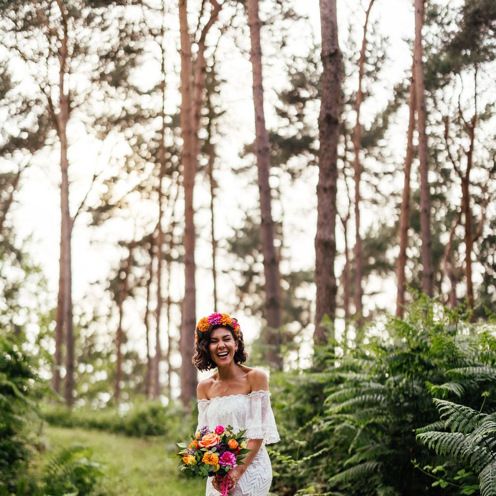 Bride in off the shoulder Grace Loves Lace wedding dress and flower crown holding colourful bridal bouquet stands in woodland after outdoor wedding ceremony at The Bridal Barn