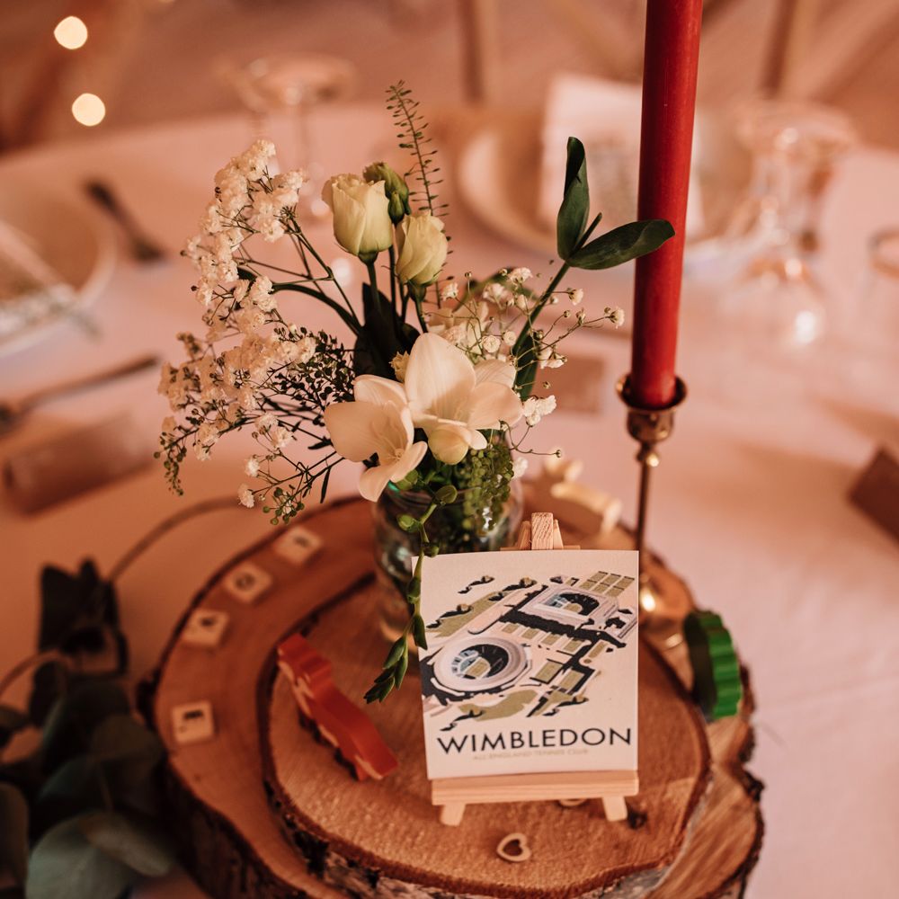 Rustic wedding centrepiece with burgundy candle, glass jar of white and green florals and Wimbledon postcard