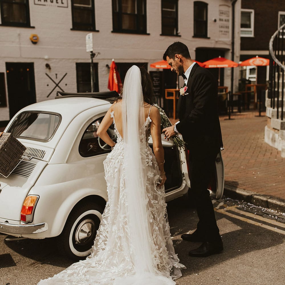 Bride wears Alena Leena Bridal gown complete with appliqué design and carries floral bouquet whilst entering vintage Fiat 500