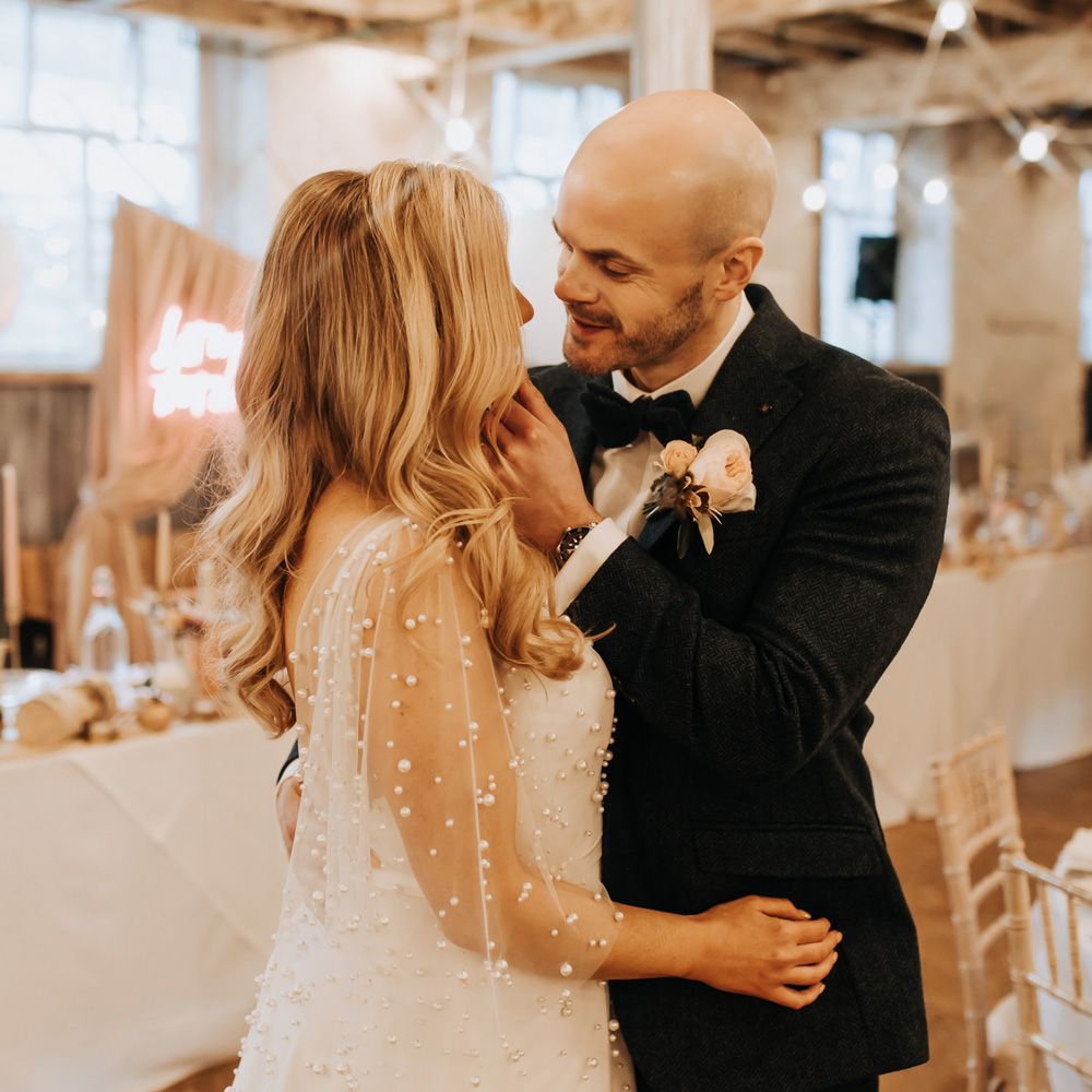 Groom in navy tweed suit with floral buttonhole stands with bride in mesh sleeve wedding dress with pearls at wedding reception