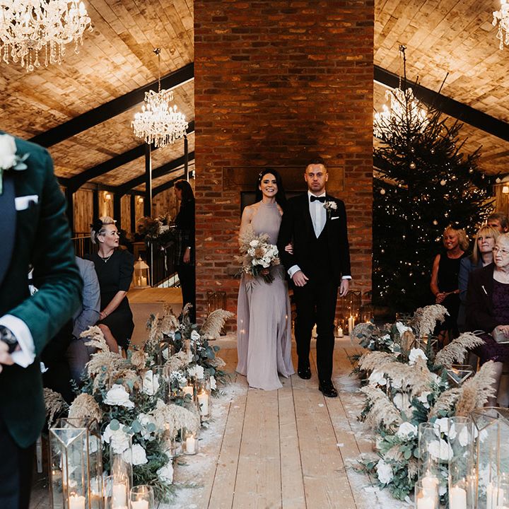 Bridesmaid in taupe dress and groomsman in black tie walk down the aisle together with chandeliers and Christmas tree decor for winter wedding 