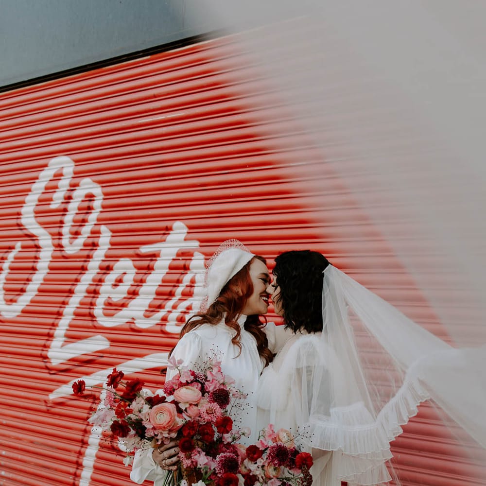Two brides kissing in a short wedding dress long veil wedding outfit and berret hat, high neck wedding dress with long sleeves holding a red and pink wedding bouquet