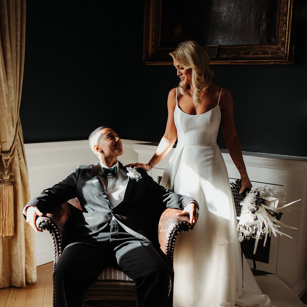 Blonde bride in a princess wedding dress with spahetti straps holding an orchid wedding boqueut next to her partner sitting in a tuxedo and bow tie 