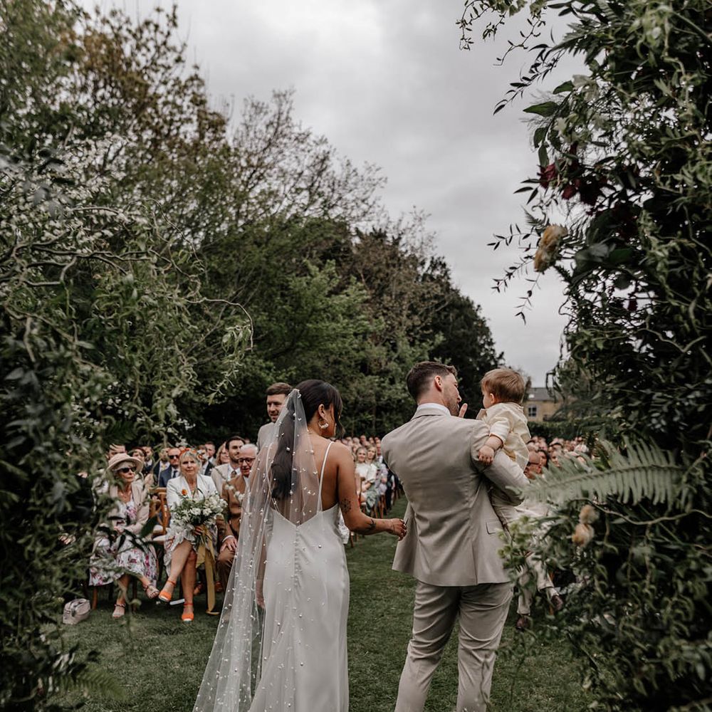 Bride in a satin wedding dress and bespoke pearl veil and groom in a beige suit holding their son at their outdoor garden wedding ceremony