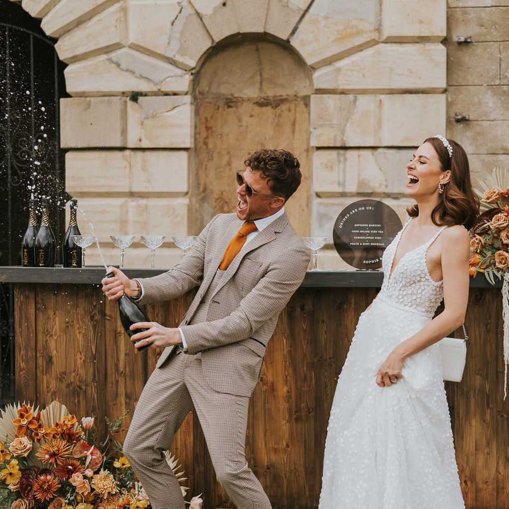 Couple in sleeveless lace wedding dress and tartan suit doing champagne pop with white confetti