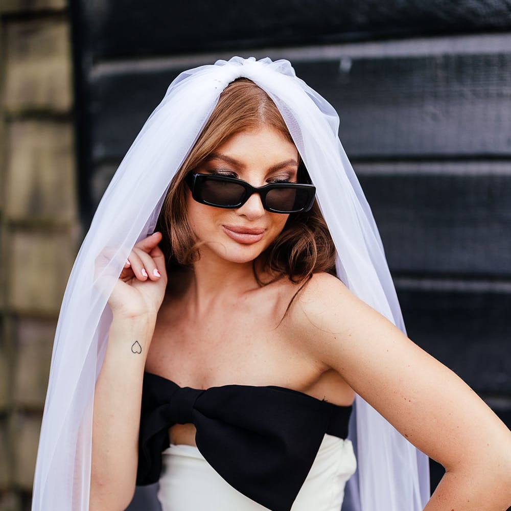 Bride wears sunglasses and cathedral veil with white mini dress complete with large black bow to front  