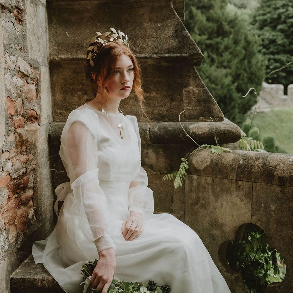 Bride with red hair sitting on a step at Bishop's Palace Wells in a two layer wedding dress and gold leaf crown holding a handed wedding bouquet 