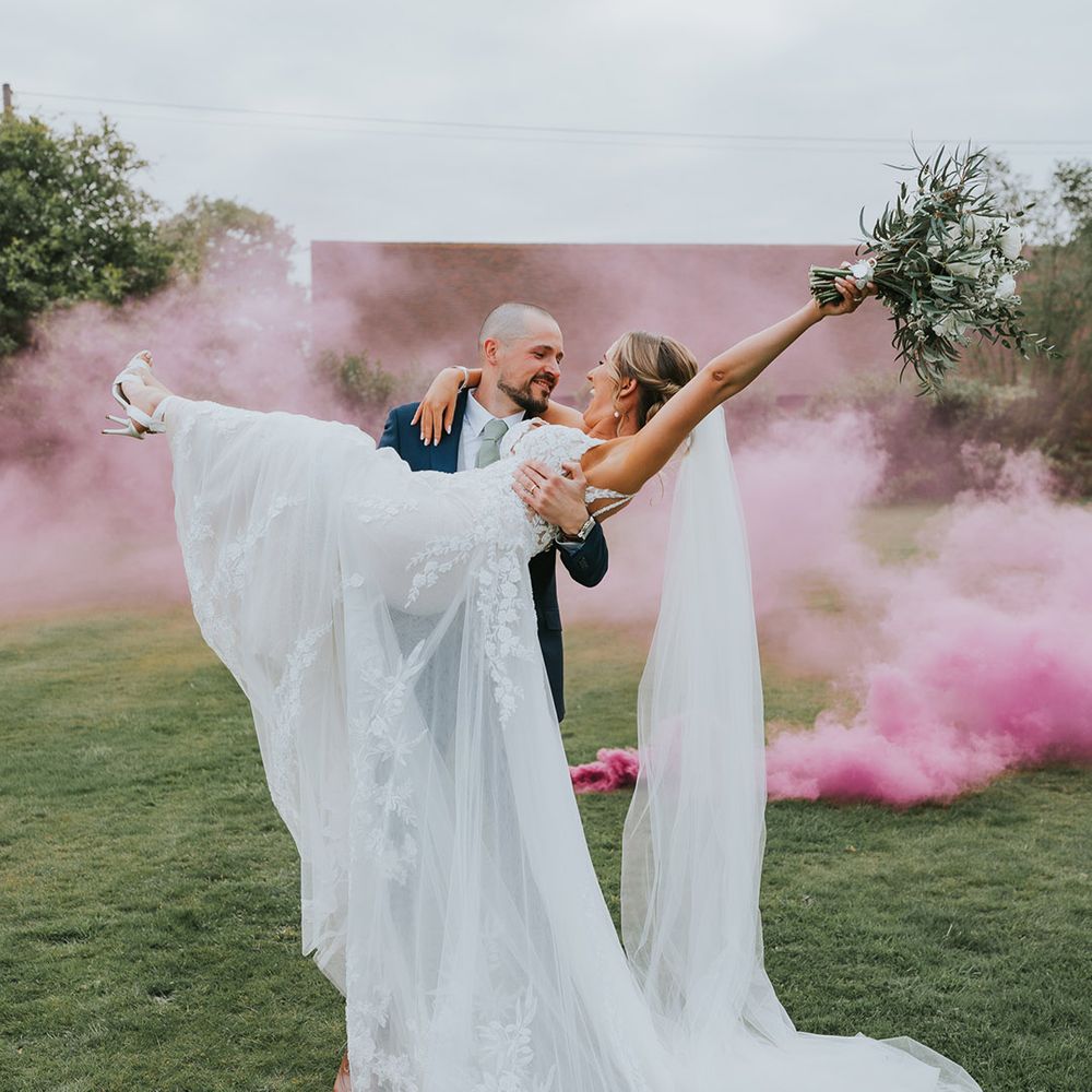 Groom in a dark blue wedding suit and bride in a lace wedding dress with a pink smoke bomb going off 