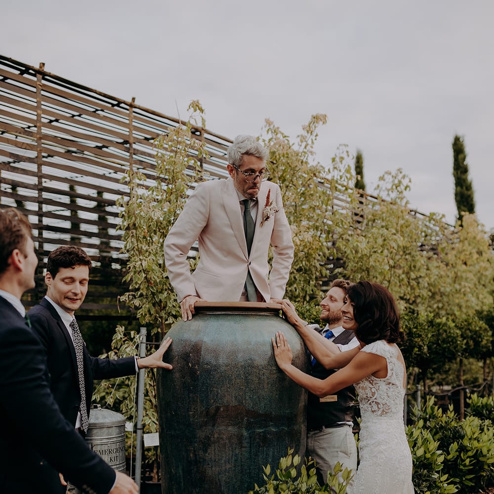 Groom gets inside a giant plant pot at Architectural Plants Venue with Bride and friends watching