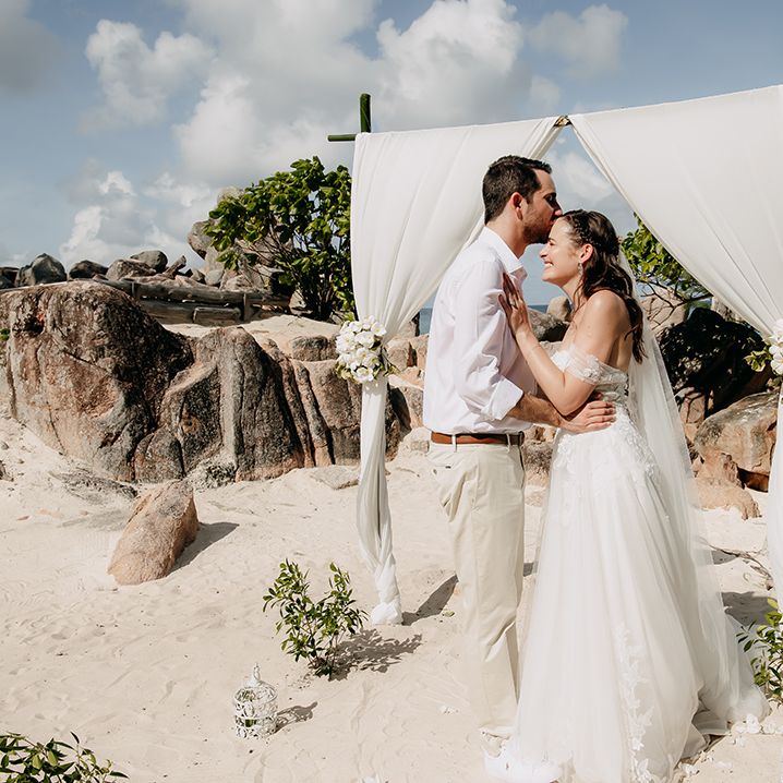 Groom in white shirt, light coloured chinos and white trainers kissing bride on the forehead wearing off the shoulder wedding dress with cathedral length veil on the beach in Seychelles