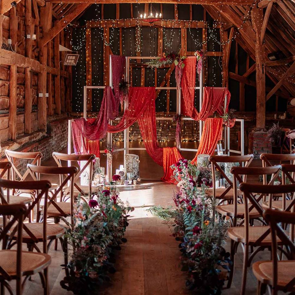 Brides standing at the end of the alter at Silchester Farm with bold crushed velvet fabrics, pheasant feathers, silver birch log stacks, candles, mismatched gold candlesticks, dried autumnal floral arrangements, red wedding drapes, fairy lights, festoon chandeliers and a string trio