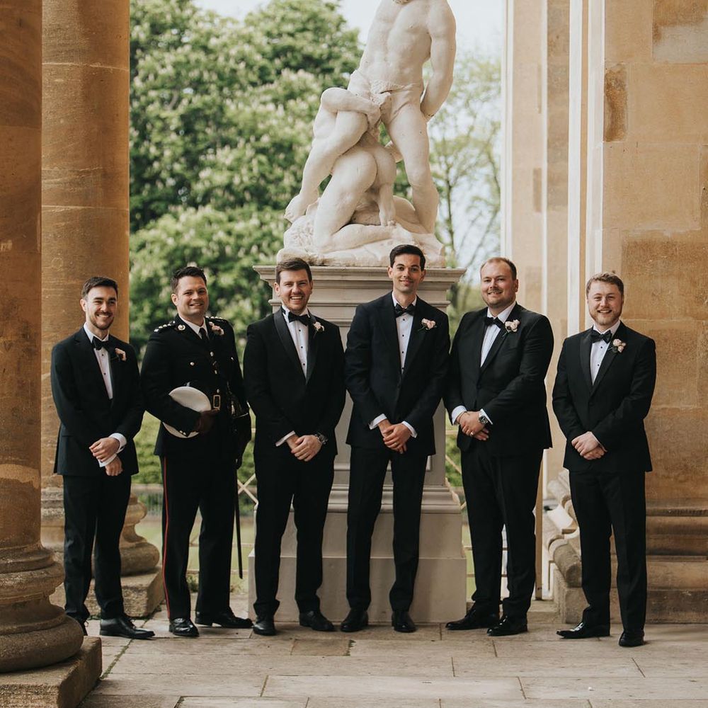 Groom and groomsmen in black tie or military uniform next to a statue at Stowe House country house wedding venue 