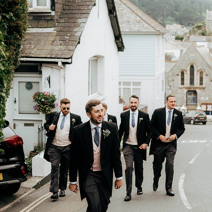 Groom & his groomsmen walk together wearing morning suits, blue ties and floral buttonholes 
