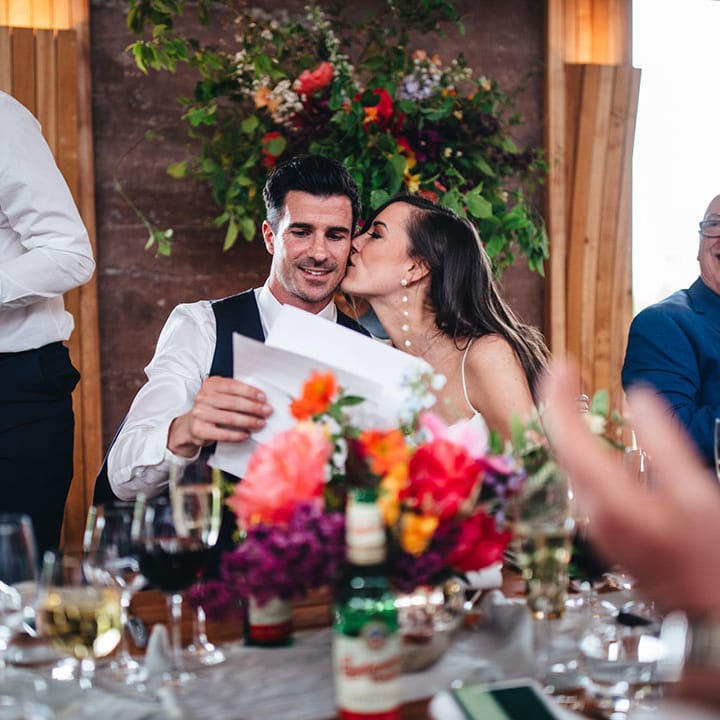 The bride wearing long statement pearl earrings leans in and kisses the groom on the cheek as he prepares to read his wedding speech 