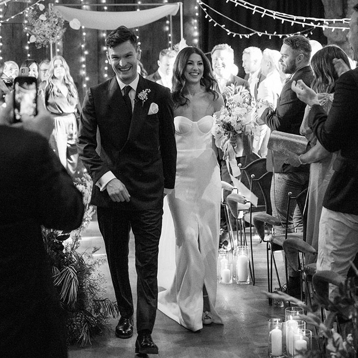 The bride and groom walk down the aisle at the rustic Tythe Barn wedding venue with white flowers 