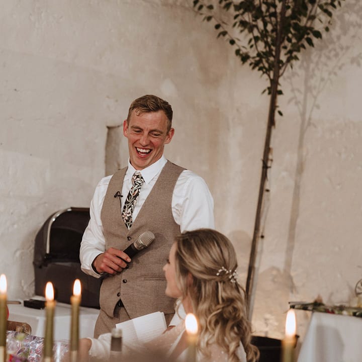 The groom in a long sleeve white shirt with a dark floral patterned tie in a beige waistcoat stand slugging as he delivers his speech 