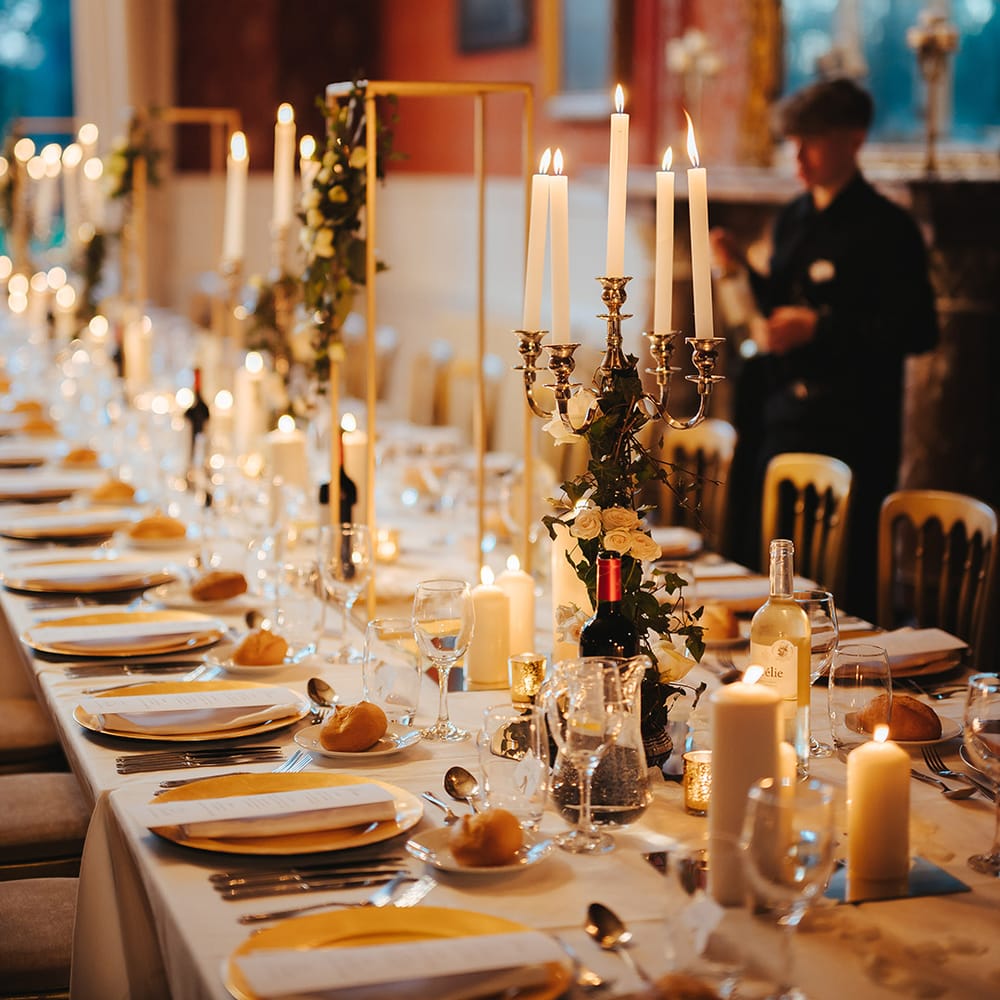 Grittleton House reception room with classic wedding tablescape including white table cloth, tapered candles in gold candelabras, pillar candles, gold frame decorations, gold plates, gold cutlery and dried flower and white rose floral arrangements 
